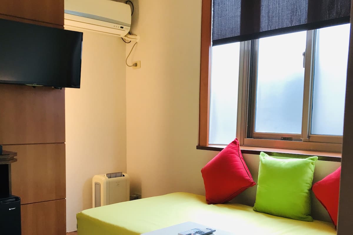 Ein kleines Zimmer mit Bett, einem kleinen Tisch, einem an der Wand befestigten Fernseher und einem Fenster mit Jalousien.
