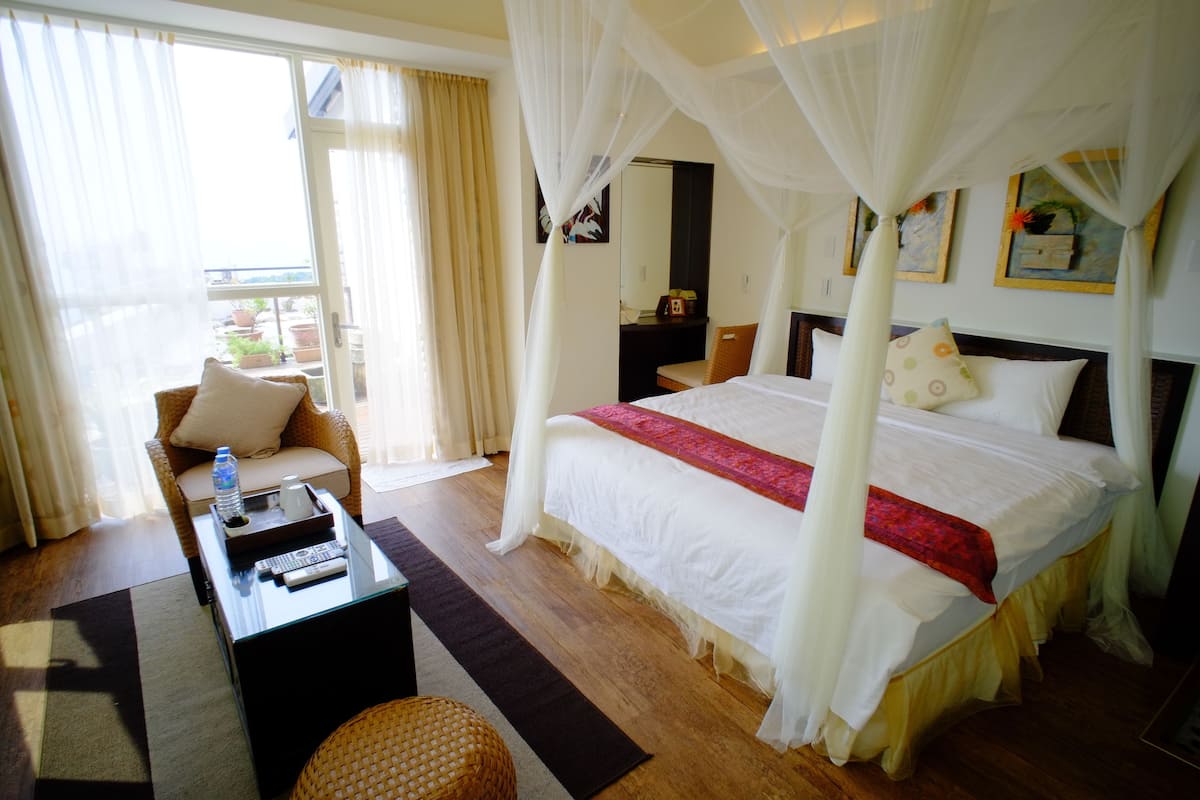 Ein Hotelzimmer mit einem großen Bett, einer Sitzecke mit Sofa und Sessel, einem Couchtisch mit Fernbedienungen und Blick ins Freie durch ein großes Fenster.