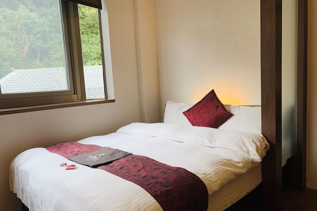 Ein ordentlich bezogenes Bett mit weißen Leinen und einem Kissen mit Rosenmuster, ein Nachttisch mit einem Buch und ein Fenster mit Blick auf Bäume.