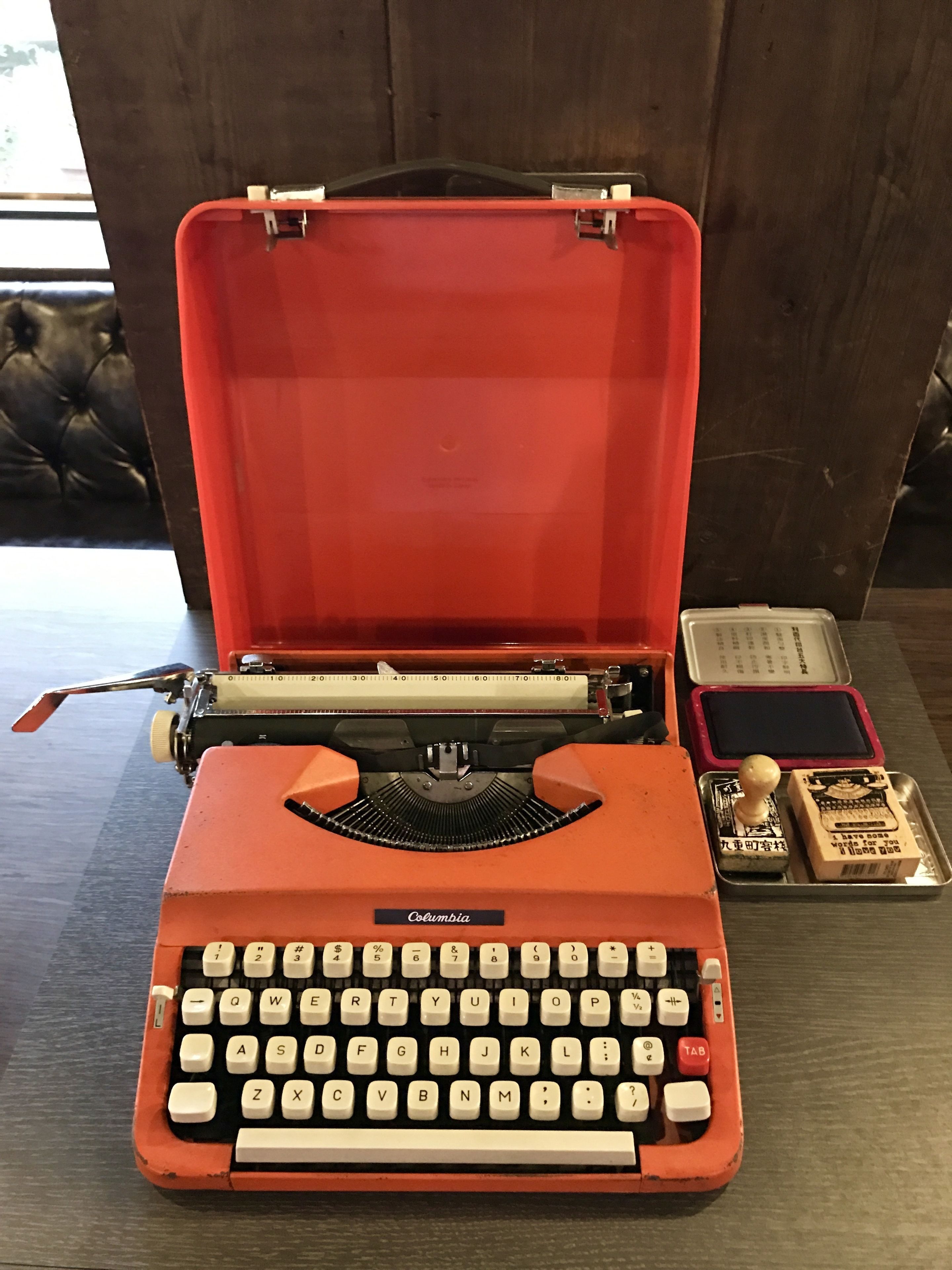 A vintage orange typewriter with a red case, a ruler, and two stamps on a wooden surface.
