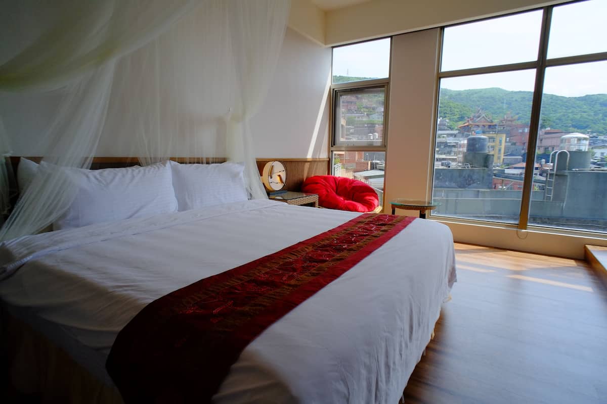 Ein Bett mit weißer Bettwäsche und einer roten Decke, ein Nachttisch mit einer Uhr und Blick auf Gebäude durch große Fenster.
