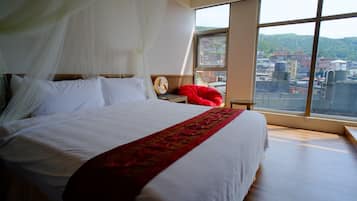 Una cama con sábanas blancas y una manta roja, una mesita de noche con un reloj, y vistas a edificios a través de amplias ventanas.