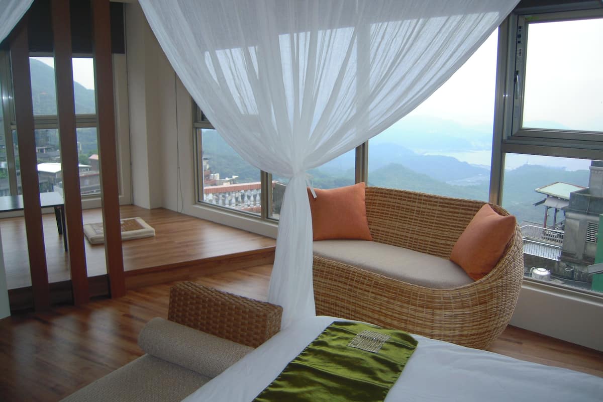 Ein Schlafzimmer mit großem Fenster, einem Korbsessel, einem Bett und Blick auf Berge.