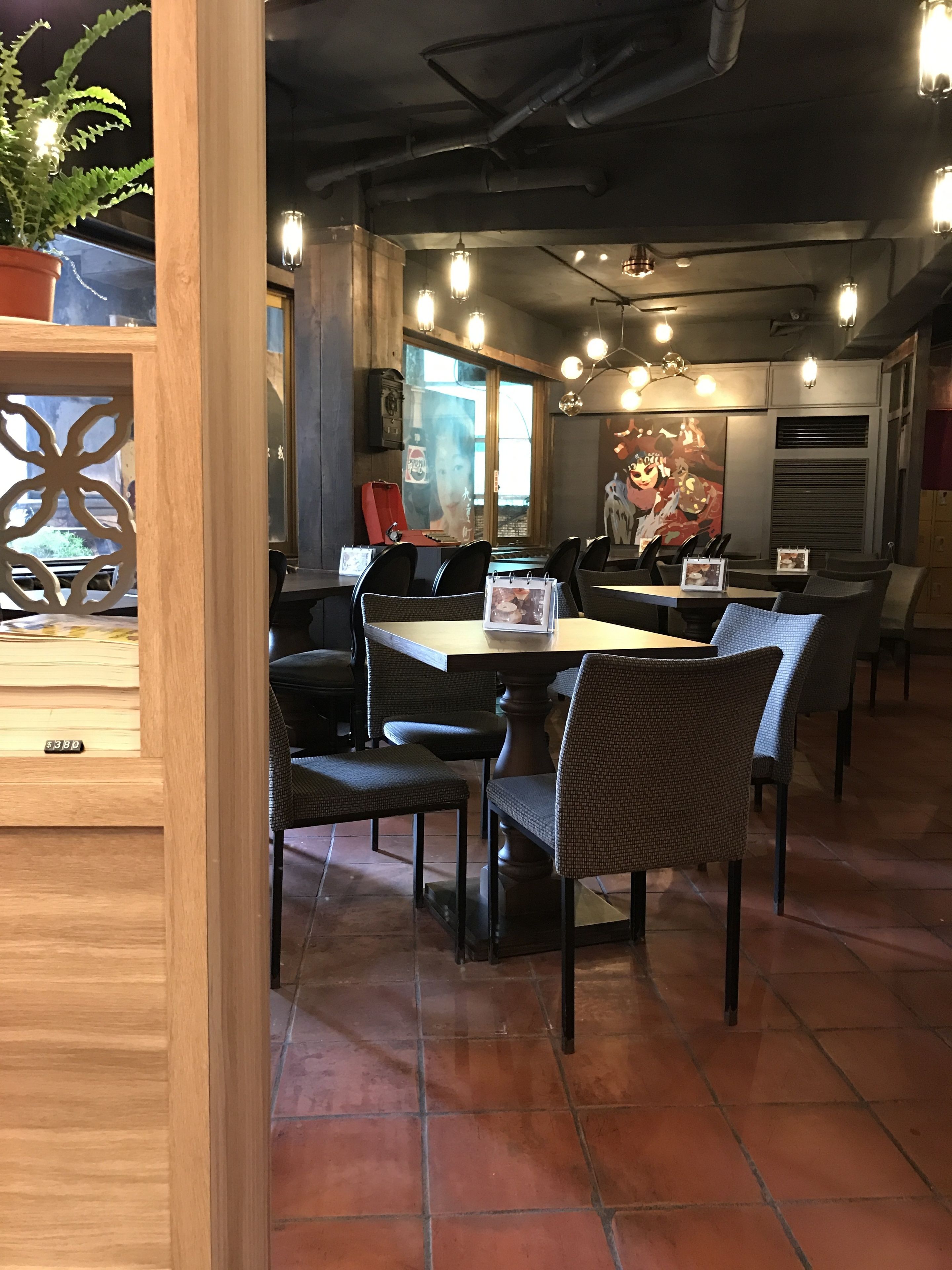A restaurant interior with tiled floors, wooden tables and chairs, pendant lighting, and a large anime poster on the wall.