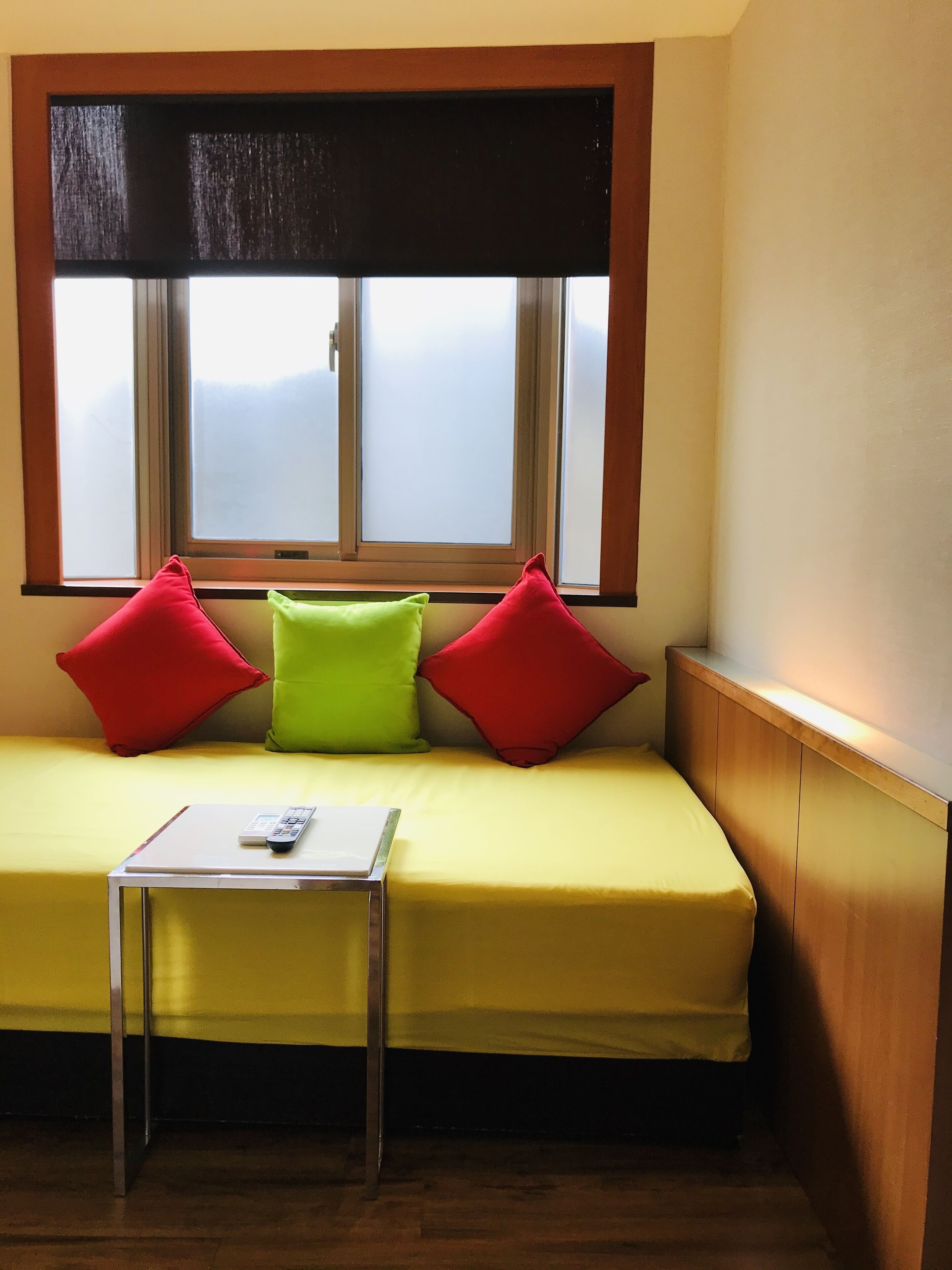 A room with a yellow sofa, three pillows, a small table with a remote control, and a window with blinds.