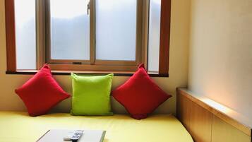 A room with a yellow sofa, three pillows, a small table with a remote control, and a window with blinds.