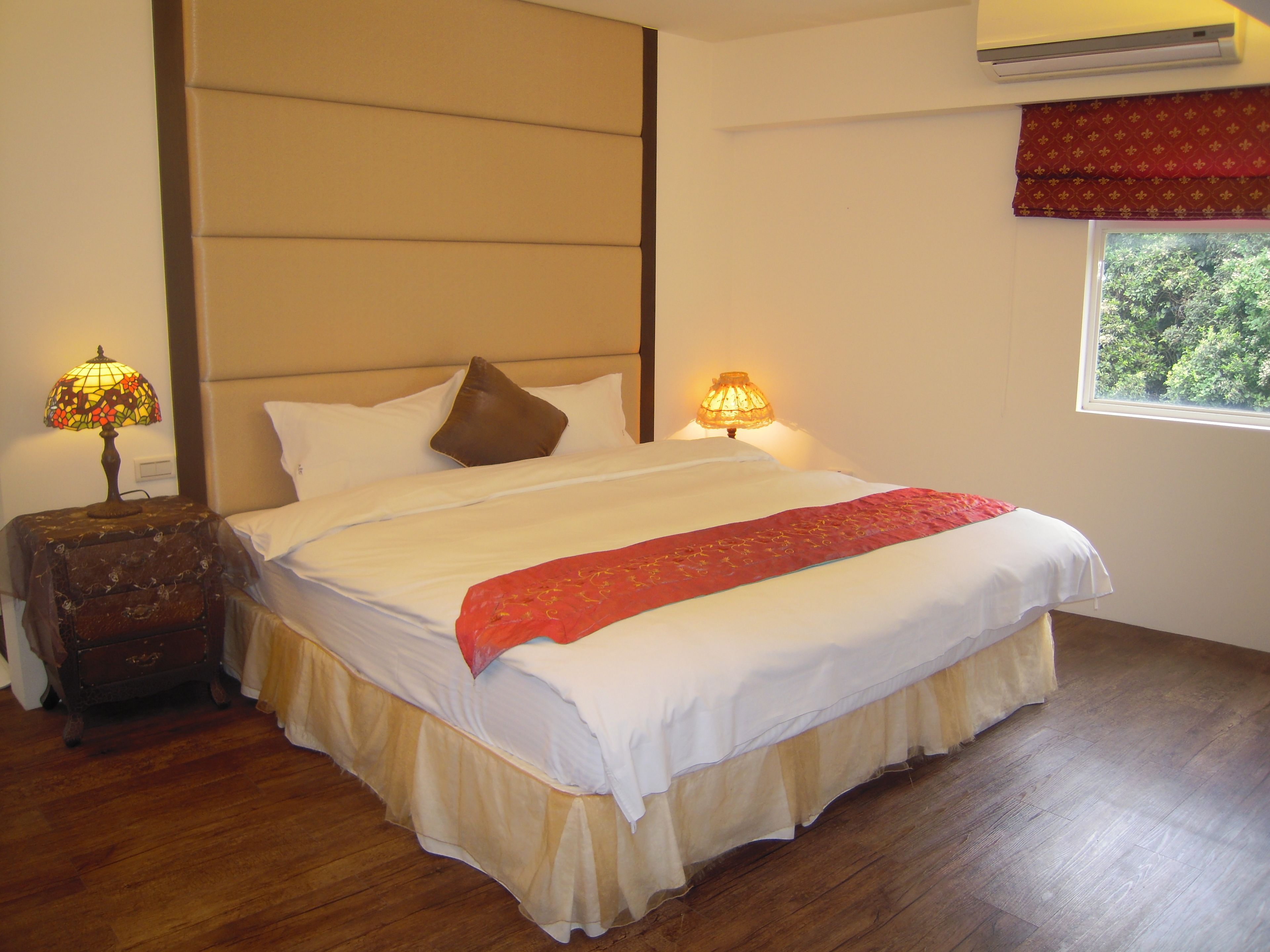 A neatly made bed with a red runner, two bedside lamps, a wooden nightstand, and a window with a view of greenery.