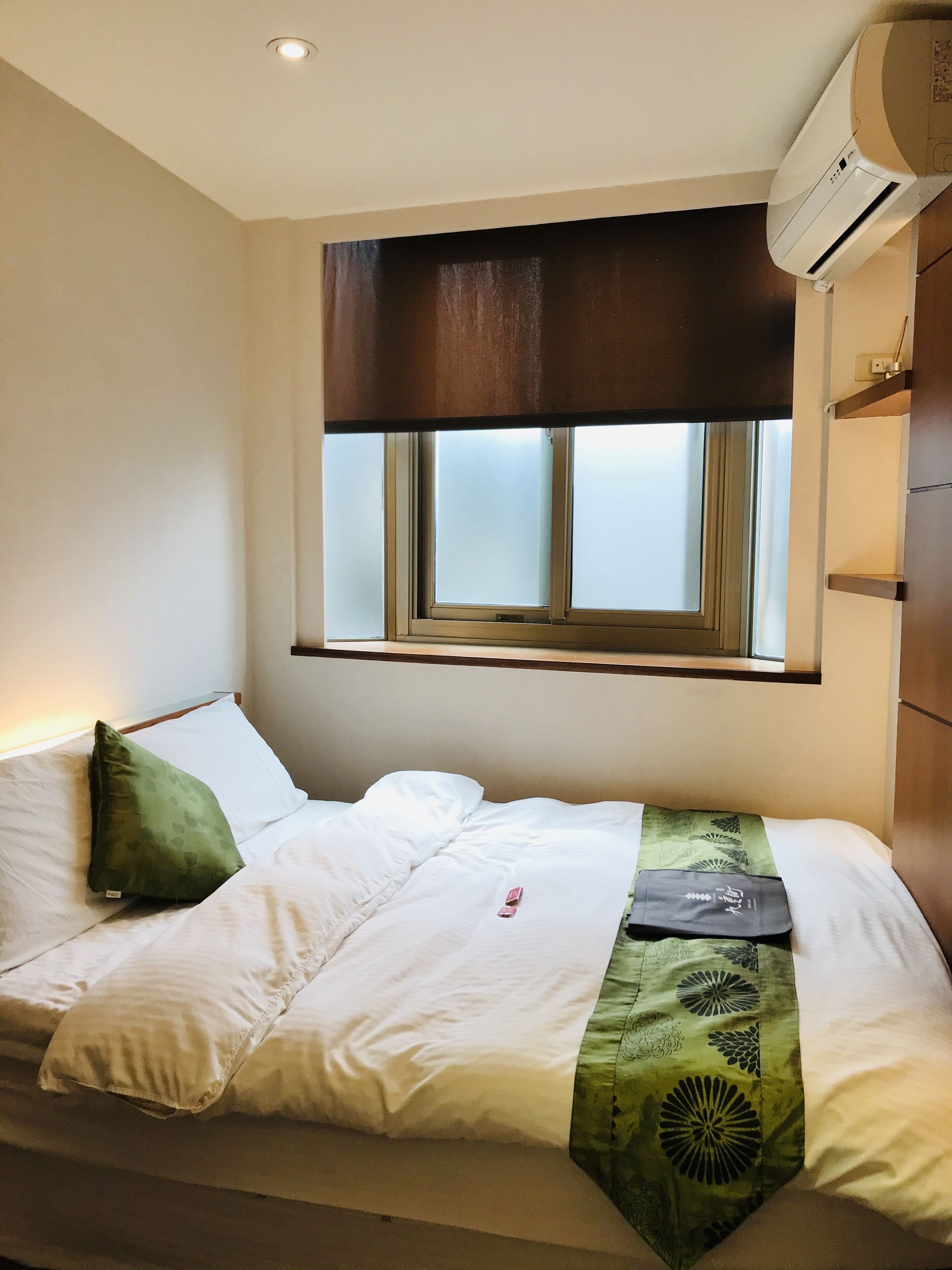 A neatly made bed with white linens and a green patterned throw, a window with blinds, and an air conditioning unit above.