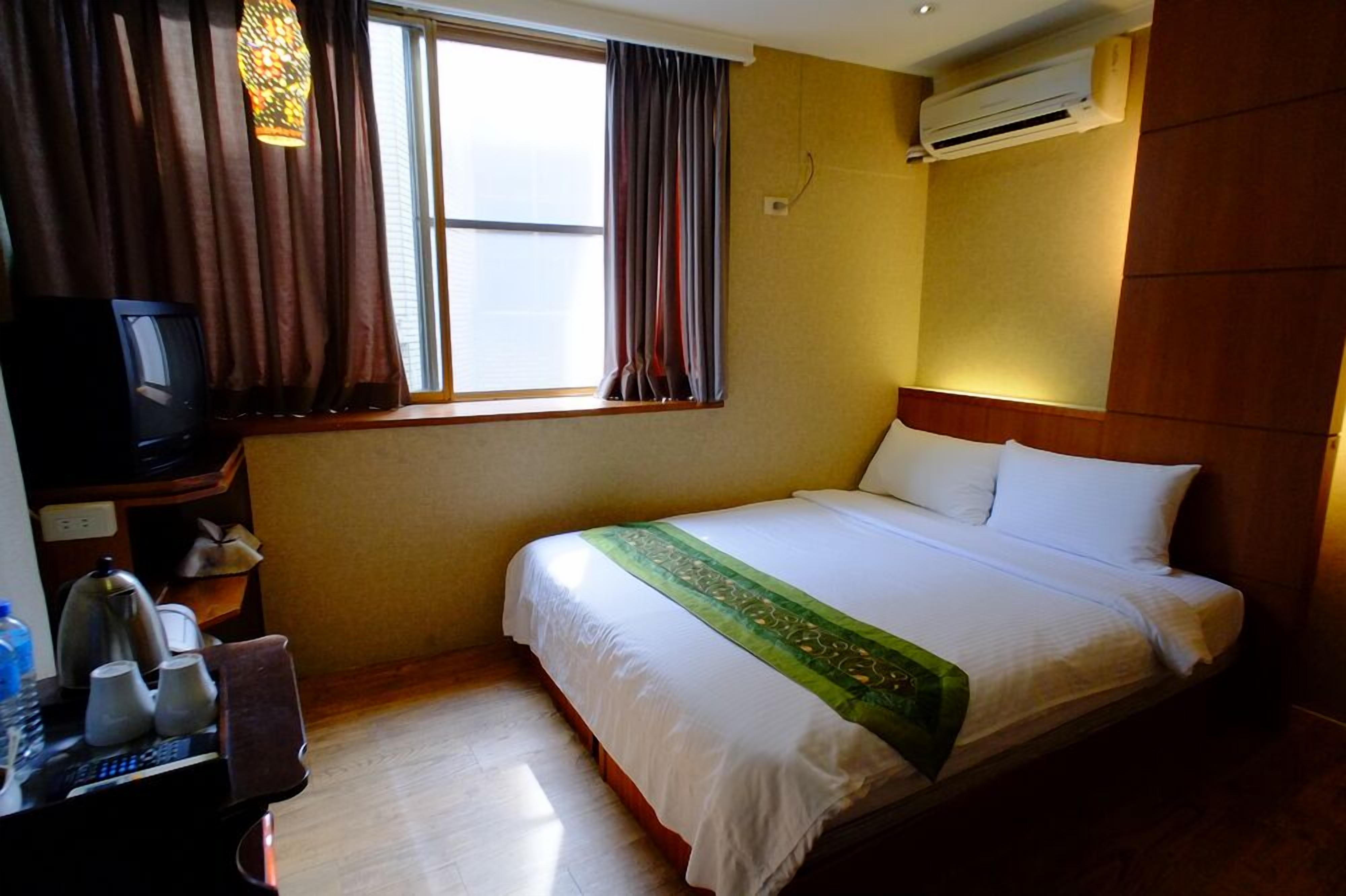 Habitación de hotel con cama, escritorio con televisor, ventana con cortinas y equipo de aire acondicionado.