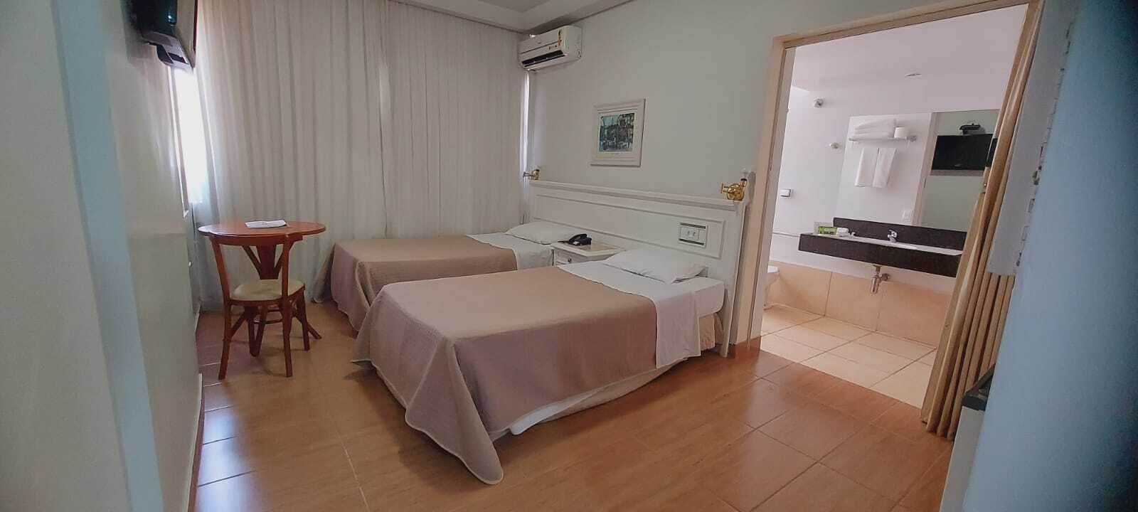 Habitación de hotel con dos camas, una mesita, una silla, una mesita de noche, un espejo y una ventana con cortinas.