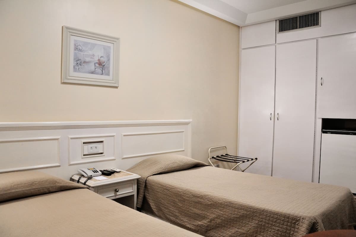 A hotel room with two beds, a bedside table with a phone and a lamp, a framed picture on the wall, and a wardrobe.