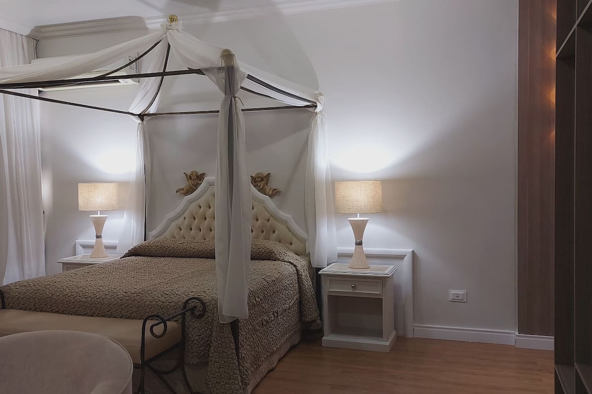 A four-poster canopy bed with a tufted headboard, two bedside tables with lamps, and a chair.