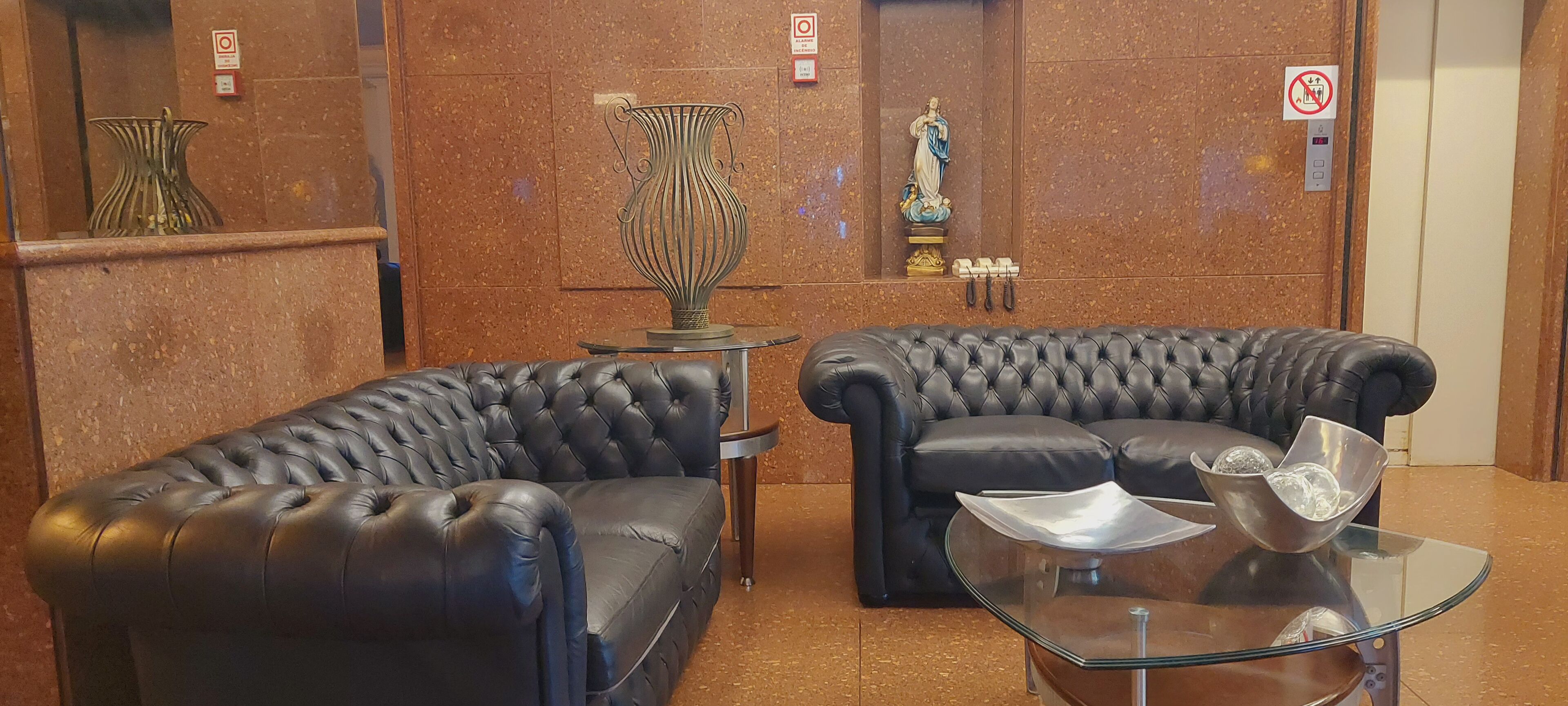 Um hall com dois sofás de couro preto, uma mesa de centro de vidro, um vaso decorativo e uma estátua na parede.