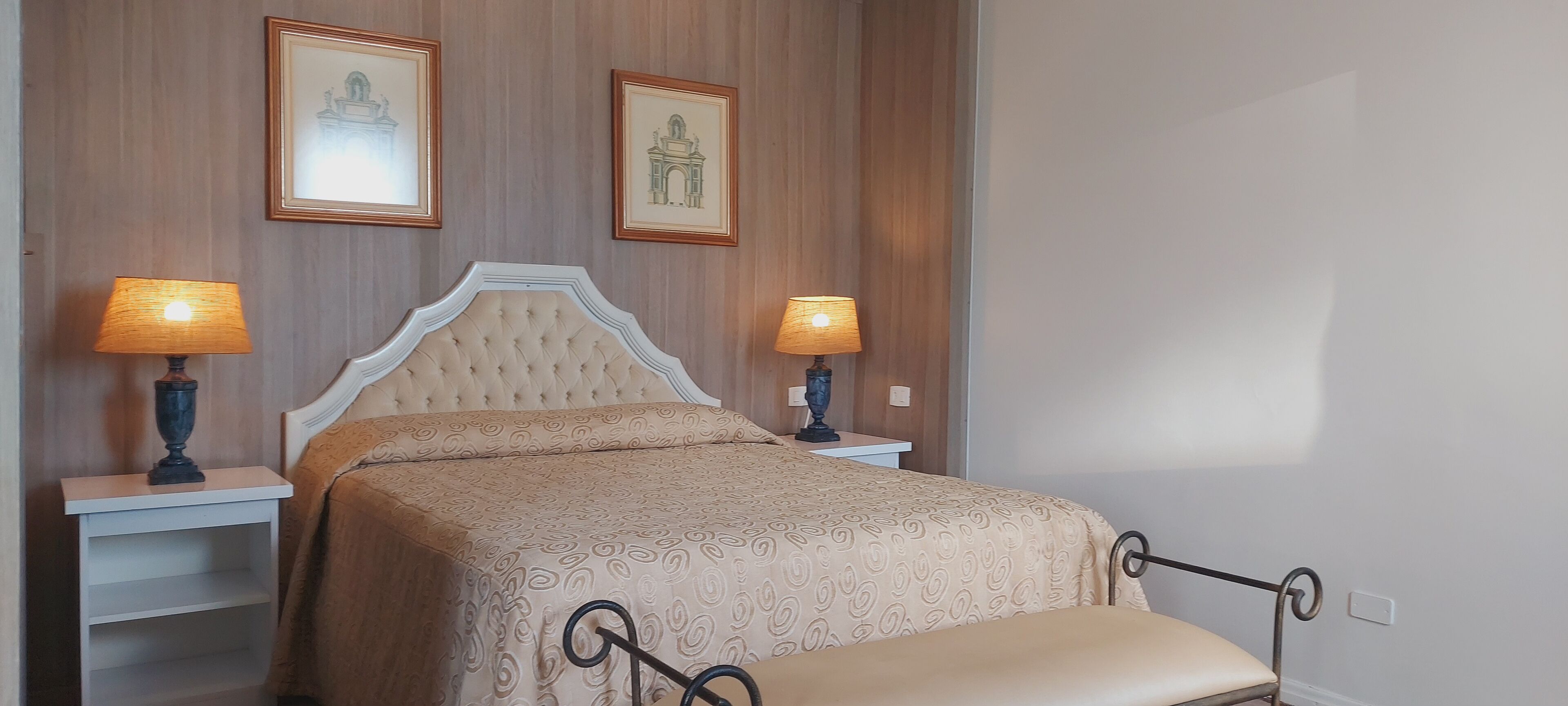 Quarto bem arrumado com cabeceira estofada, duas mesas de cabeceira com luminárias e um banco no pé da cama.