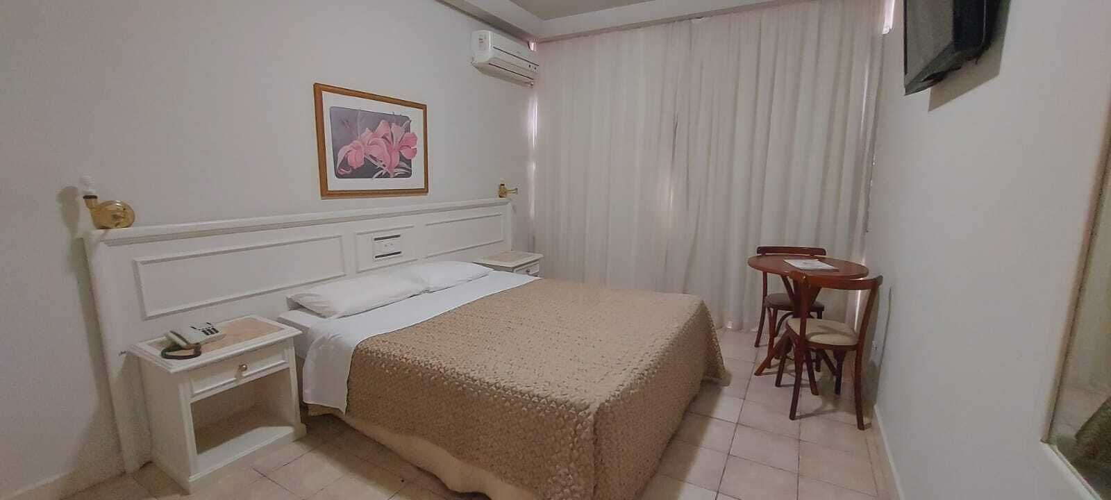 Quarto de hotel com cama, mesa de cabeceira, cadeira e uma pequena mesa com outra cadeira.
