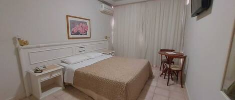 Quarto de hotel com cama, mesa de cabeceira, cadeira e uma pequena mesa com outra cadeira.