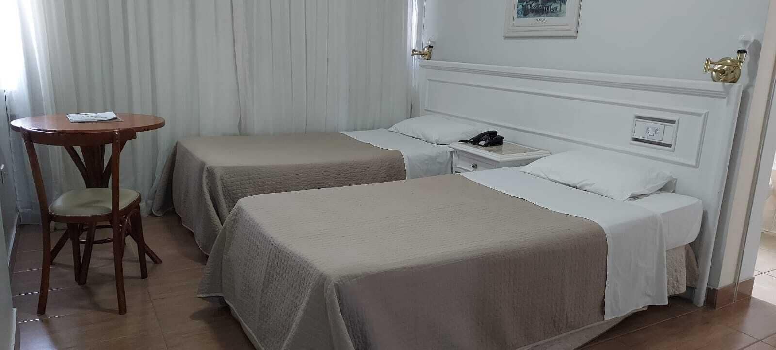 A hotel room with two single beds, a small table with a chair, a bedside table with a phone, and a wall-mounted light.