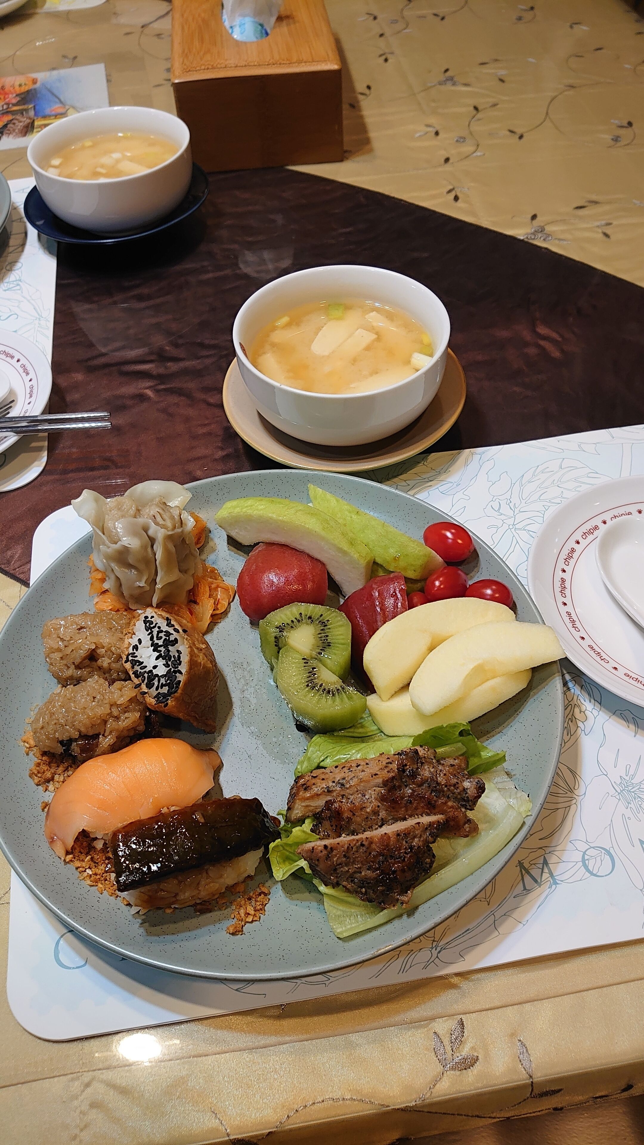 Een eettafel met diverse gerechten, waaronder soep, dumplings, sushi en gegrild vlees.
