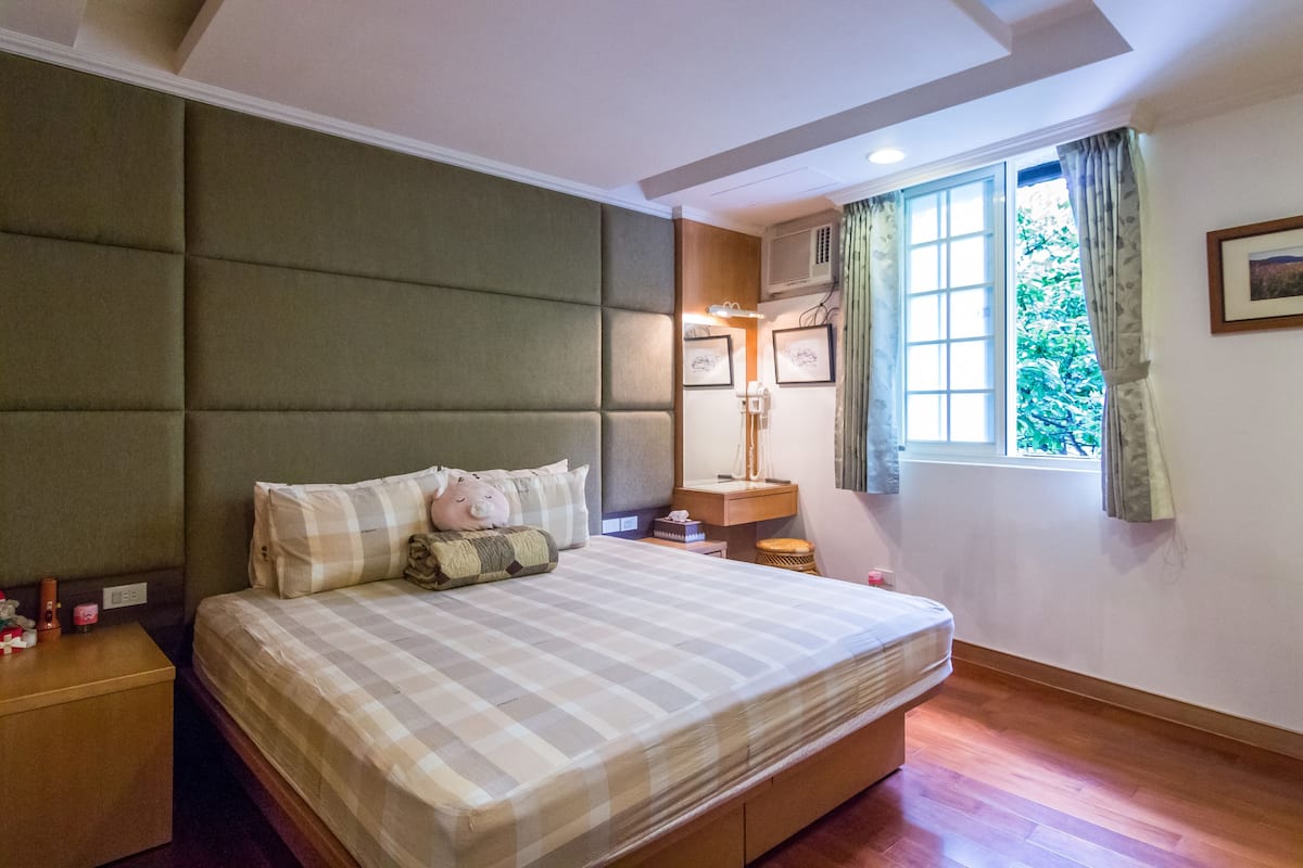 Ein ordentlich eingerichtetes Schlafzimmer mit einem großen Bett, Nachttischen, einer Nachttischlampe, einem Fenster mit Vorhängen und einer Wand-Klimaanlage.