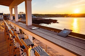 Una terraza de madera con mobiliario exterior, con vistas a un cuerpo de agua y una costa rocosa.