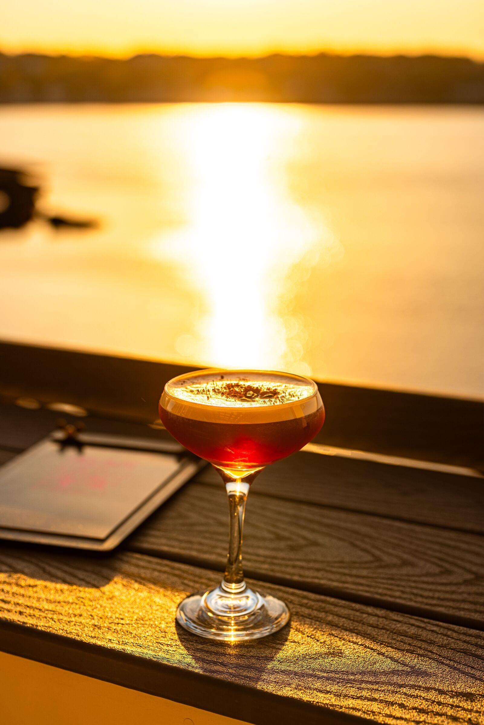 Ein Cocktailglas auf einer Holztischplatte, im Hintergrund ein Sonnenuntergang.