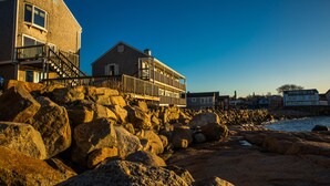 A coastal building with a balcony, large rocks in the foreground, and a clear sky. - Bearskin Neck Motor Lodge (Rockport)
