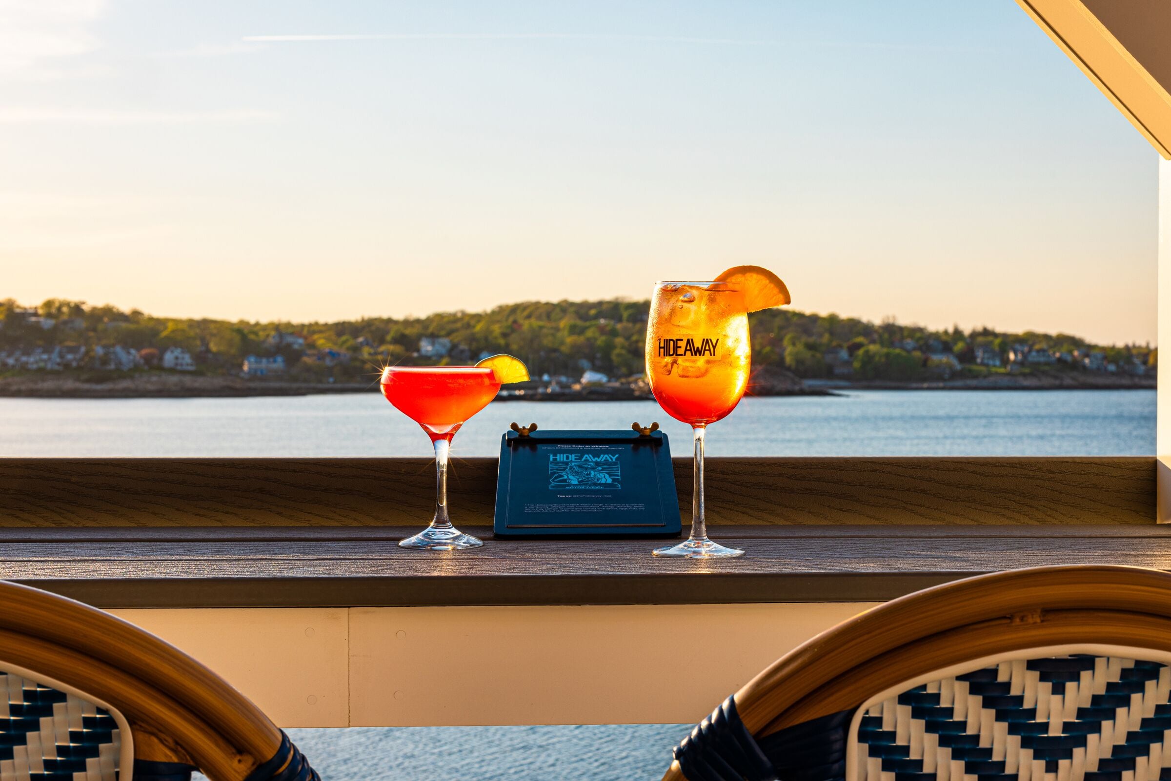 Two cocktails on a wooden bar counter with a view of a body of water and a forested area in the background.