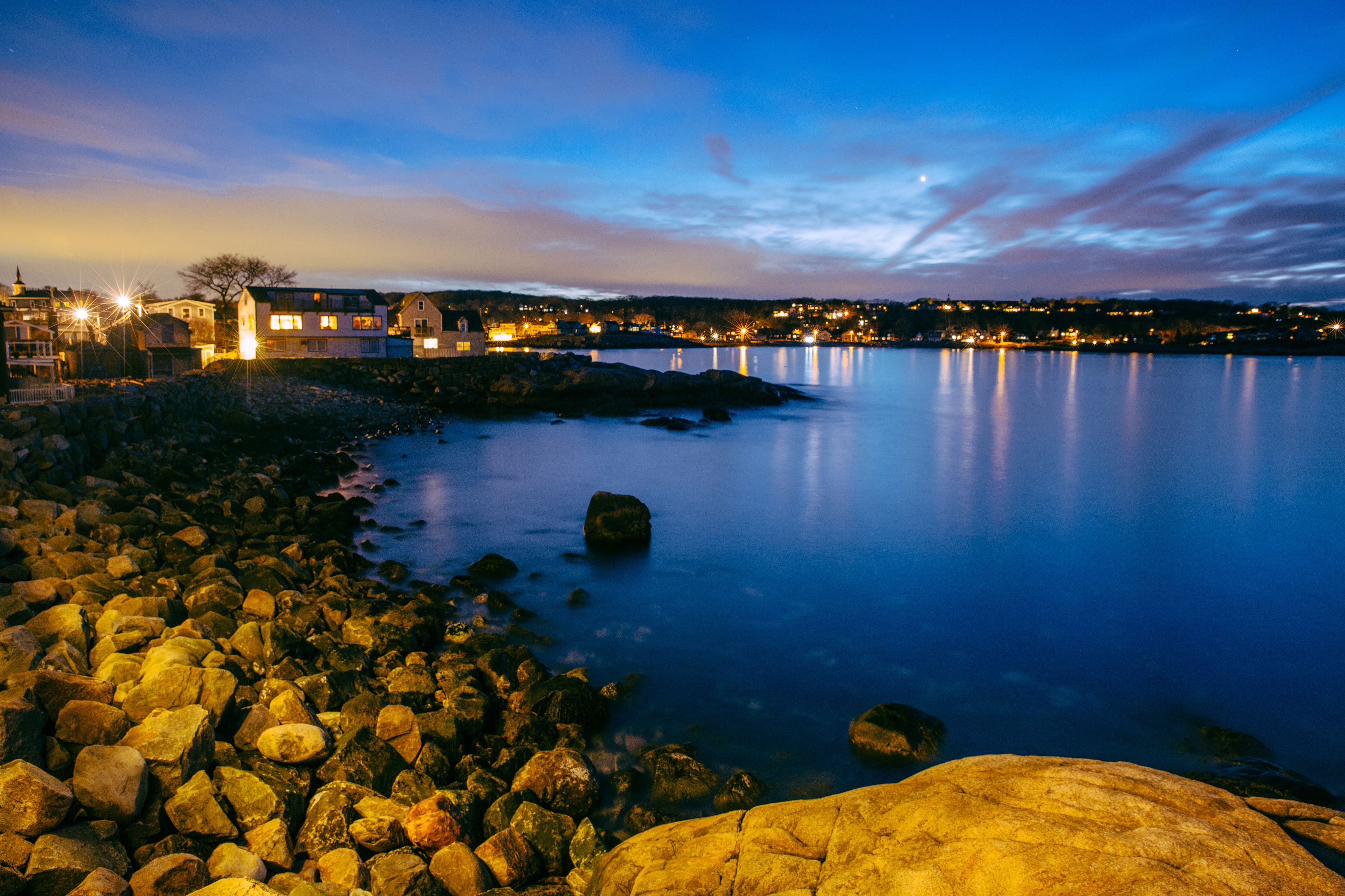 Un paysage côtier avec une rive rocheuse, une eau calme et une ville dont les bâtiments sont illuminés en arrière-plan.