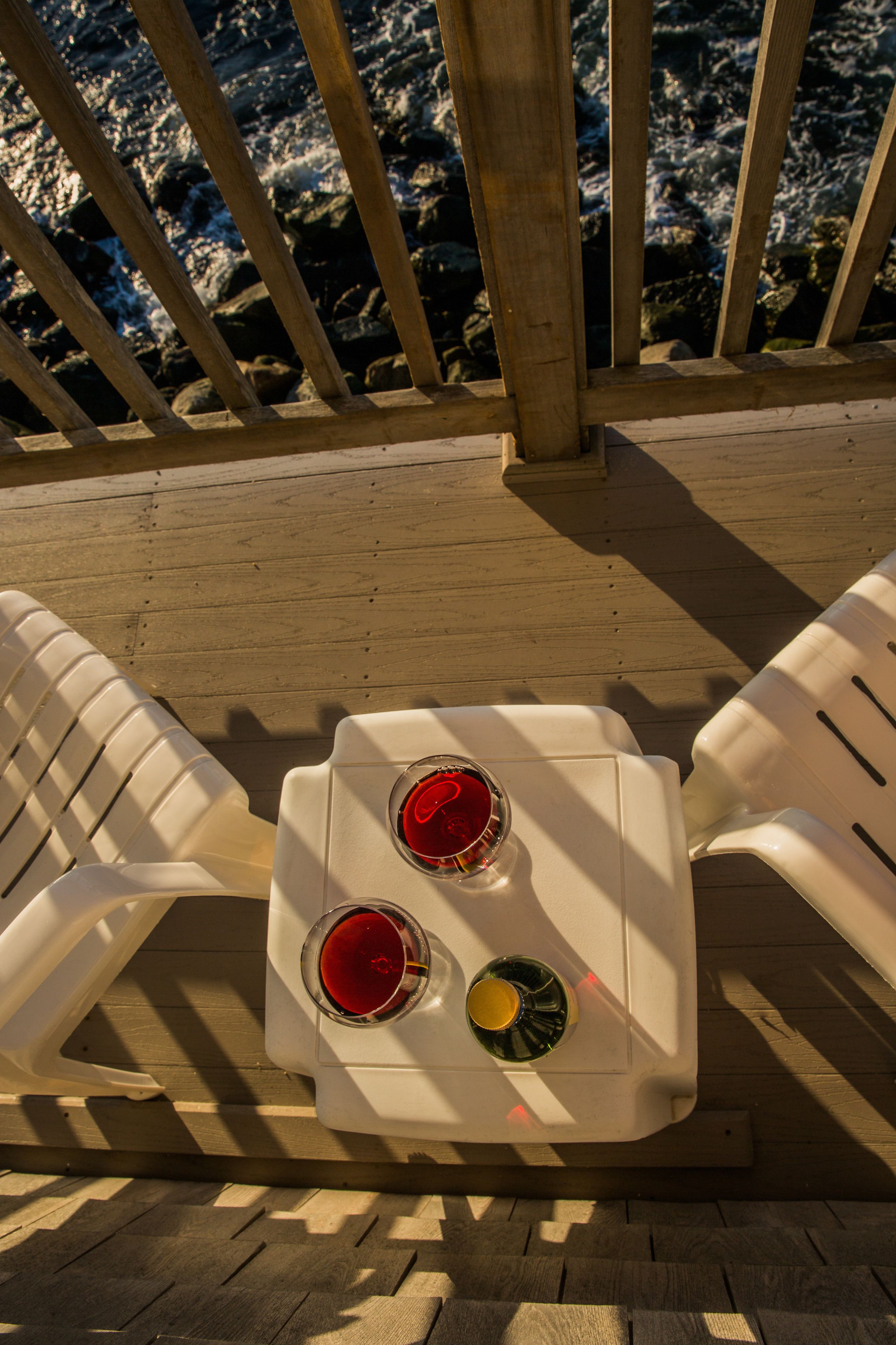 Una terraza de madera con dos sillas de plástico blanco y una mesa pequeña con dos vasos de líquido rojo y uno de líquido amarillo.