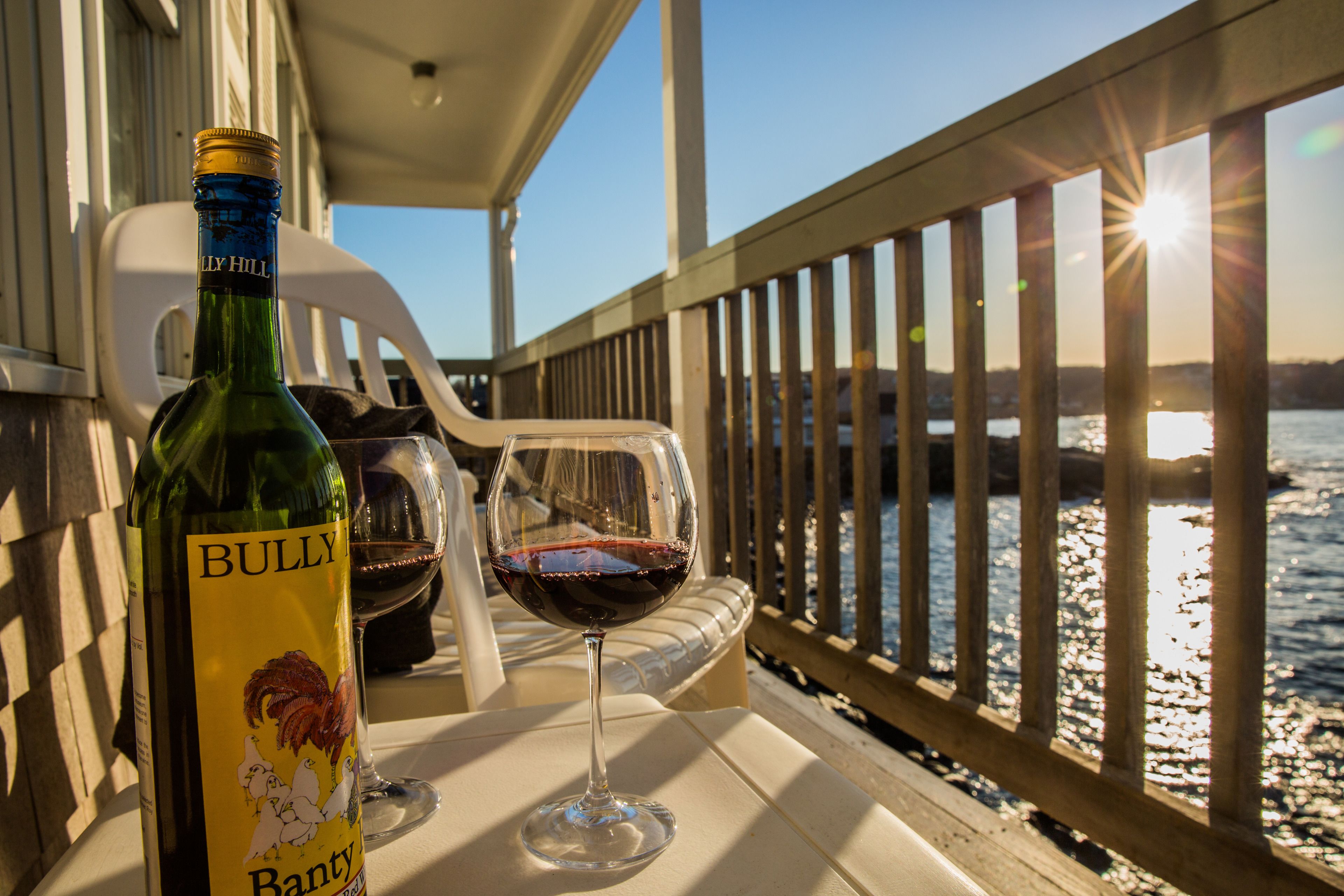 A bottle of BULLY wine and a glass on a table by a railing with a view of a body of water and the sun setting.