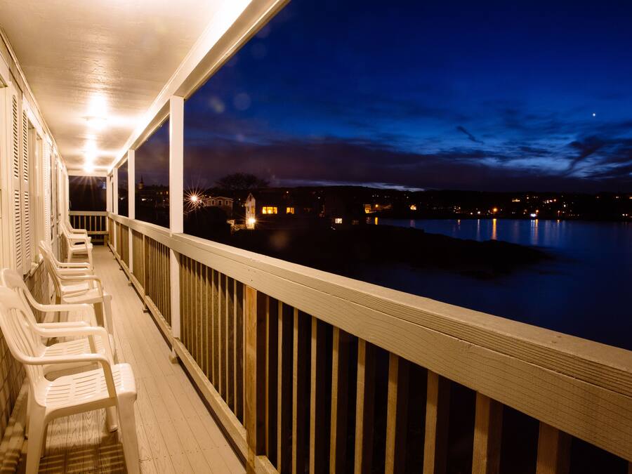 A wooden deck with white chairs and a railing overlooking a calm body of water at night.