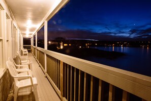 Eine Holzterrasse mit weißen Stühlen und Geländer, die auf ruhiges Wasser bei Nacht blickt.