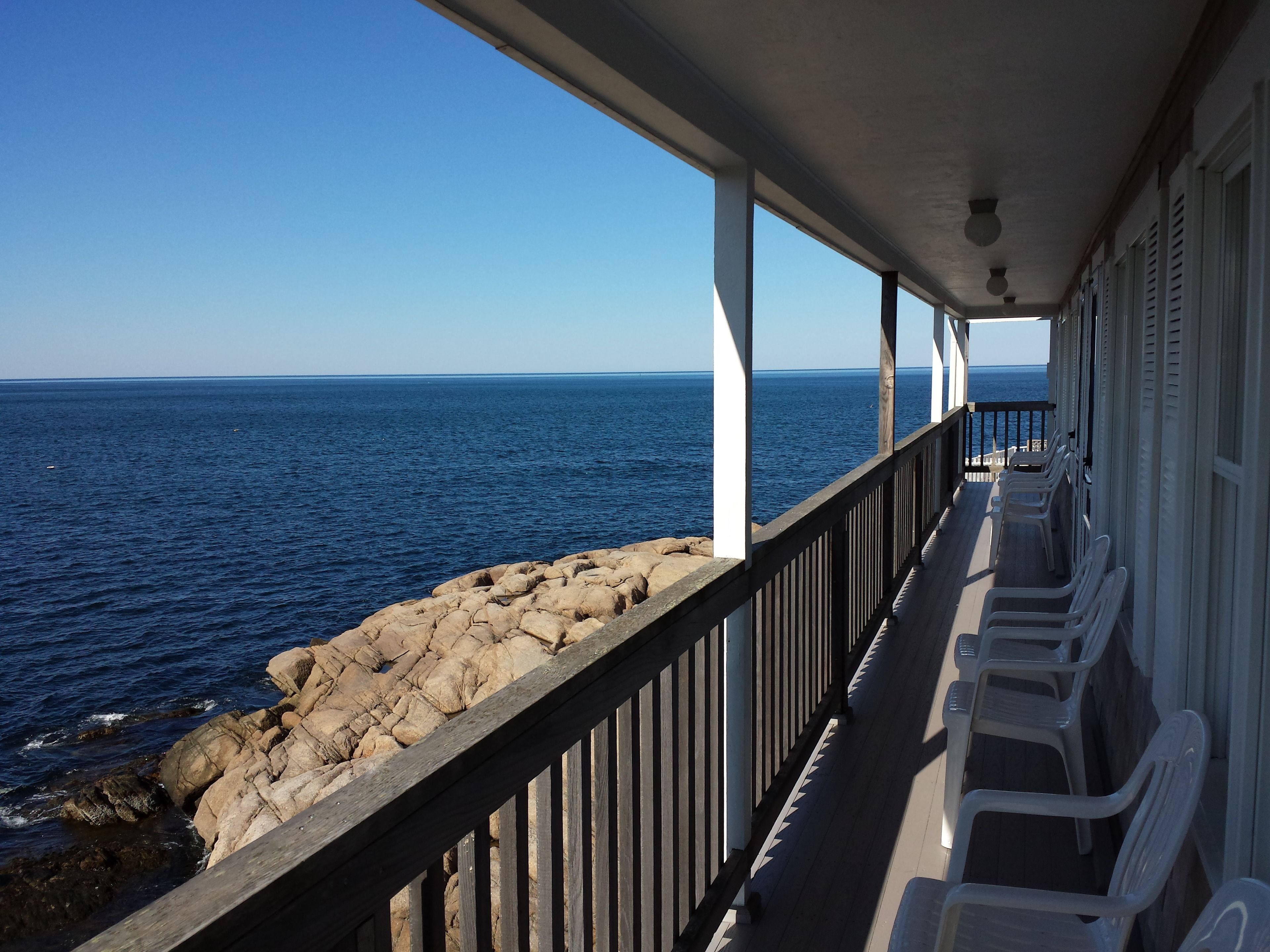 A covered outdoor area with a view of the ocean, featuring a wooden railing, white chairs, and a stone wall.