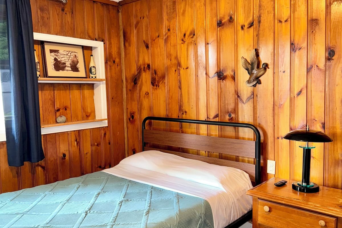 A bedroom with a wooden paneled wall, a bed with a quilt, a bedside table with a lamp, and a shelf with a framed picture and a bottle.