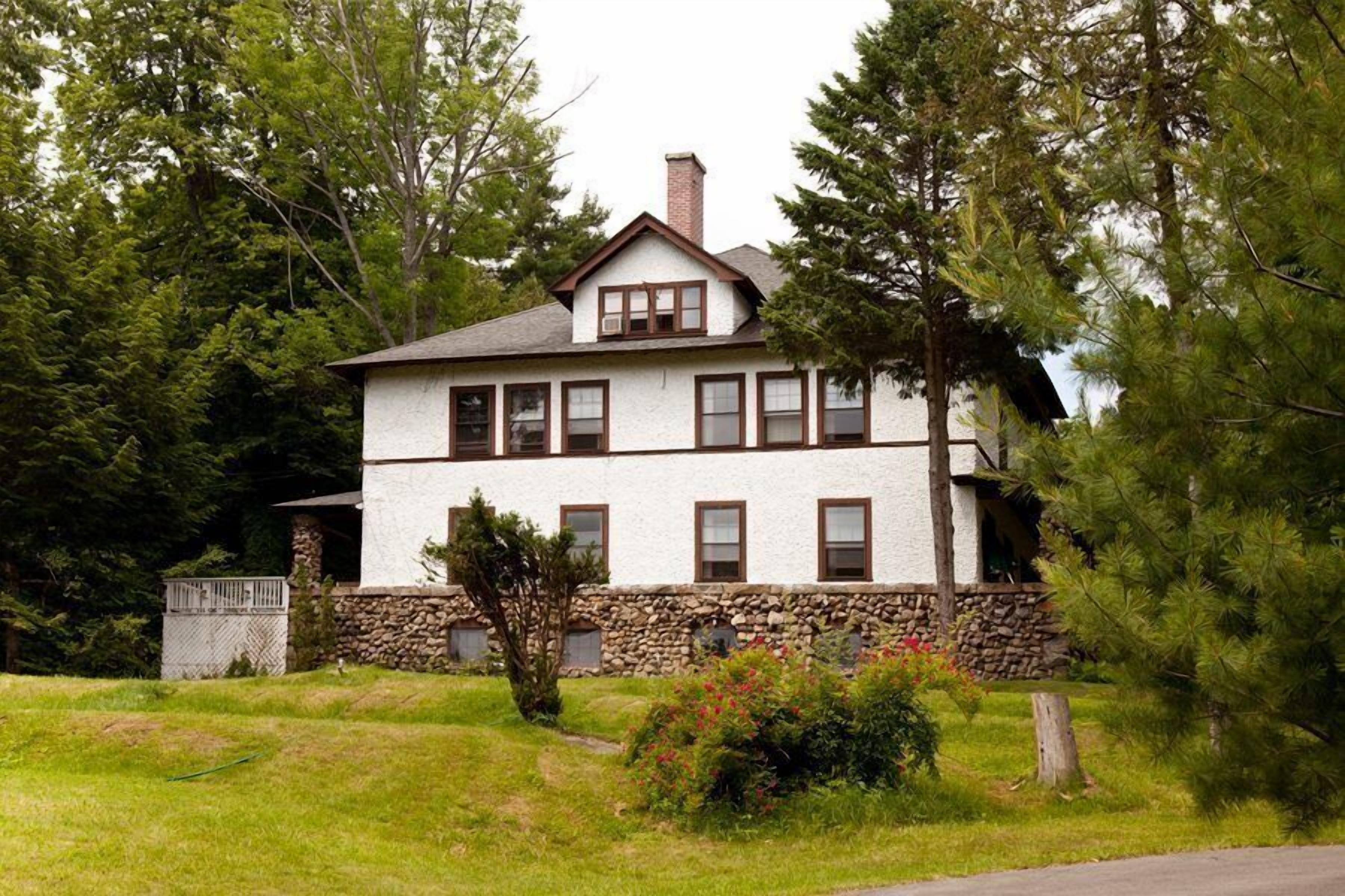 Une maison à deux étages, avec des fondations en pierre et des murs extérieurs blancs, entourée d’une pelouse et d’arbres.