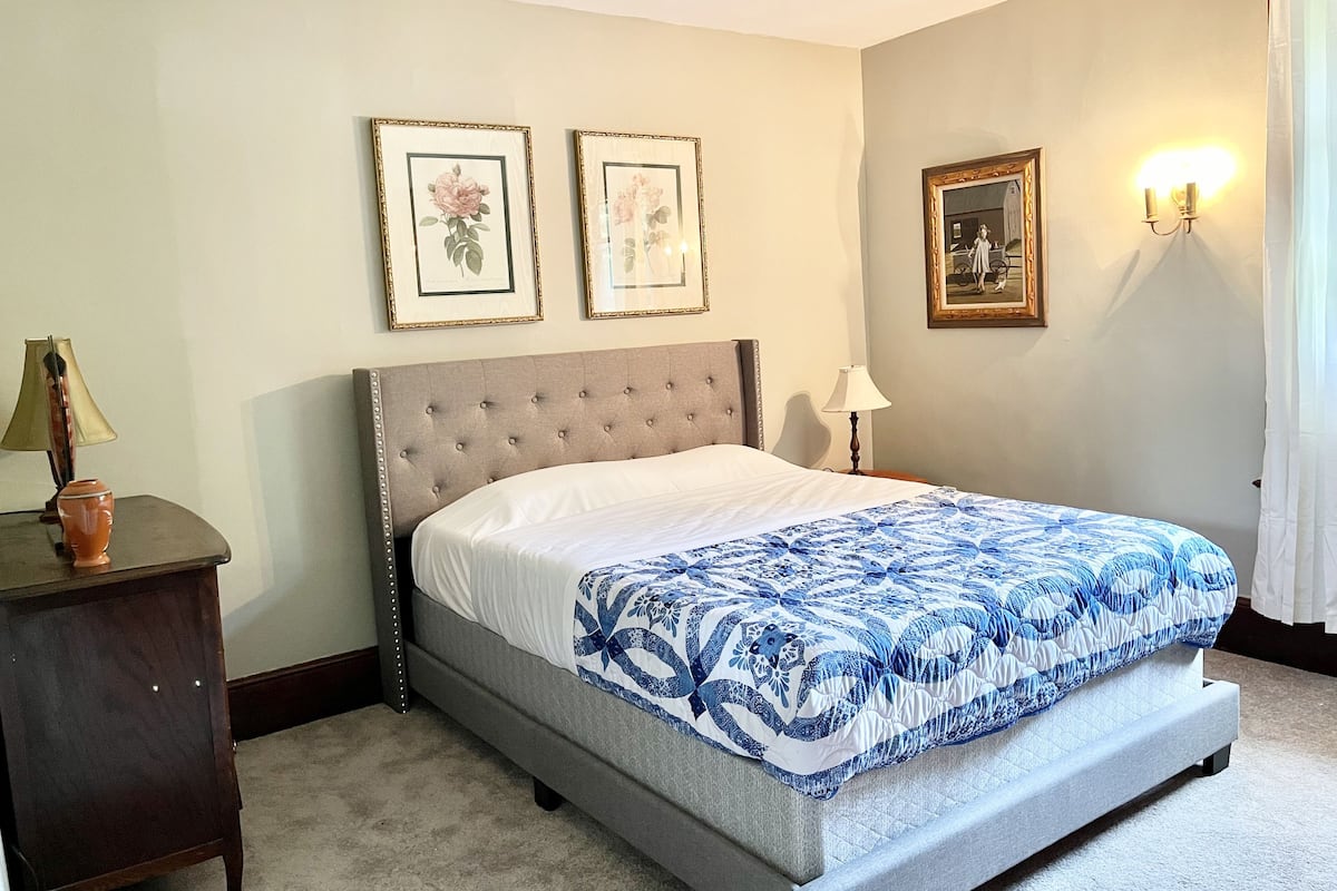 A bedroom with a bed, two framed pictures on the wall, a nightstand with a lamp, and a wall-mounted light fixture.