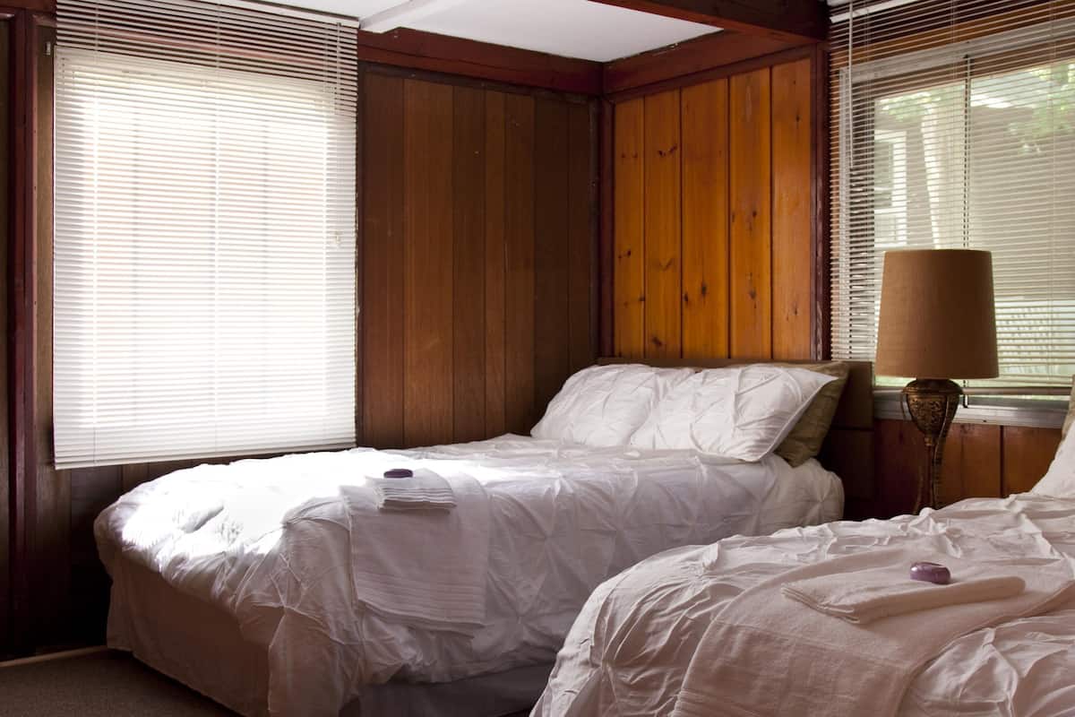 A bedroom with two beds, wooden paneling on the walls, and a lamp on the nightstand.