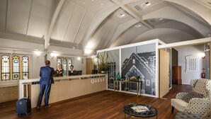 A reception area with a wooden floor, a reception desk labeled "QUEST," a seating area with patterned furniture, and a large window with stained glass. - Quest East Perth (East Perth)