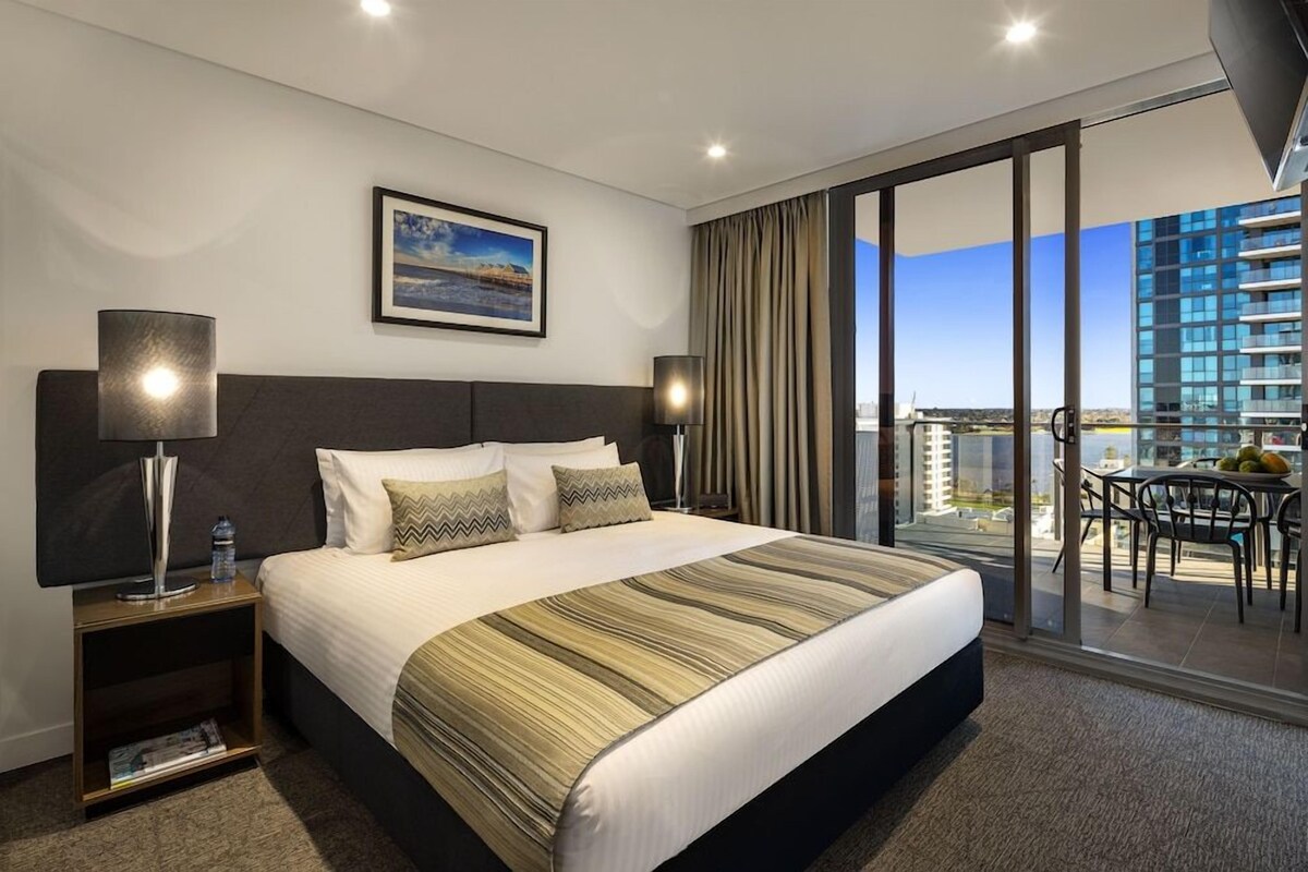A hotel room with a large bed, two bedside lamps, a framed picture on the wall, and a balcony with a table and chairs.