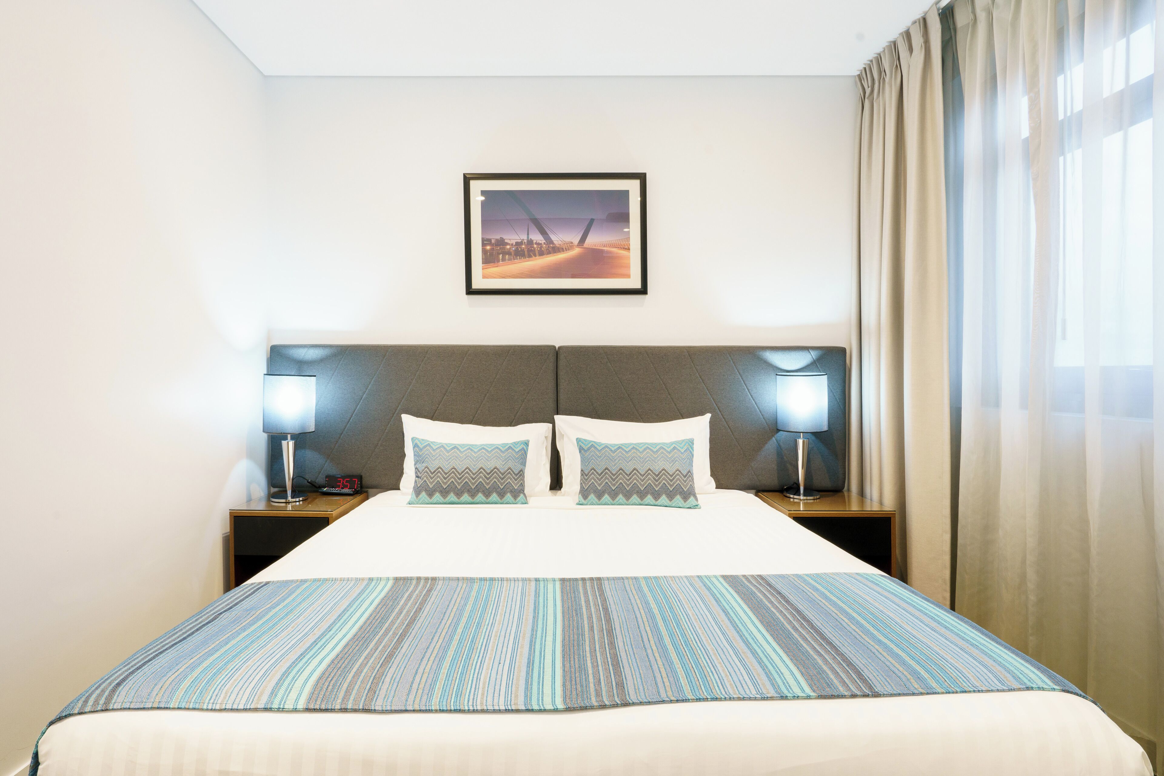 Une chambre d’hôtel avec un lit, deux lampes de chevet, une table de nuit, un tableau encadré au mur et une fenêtre avec des rideaux.