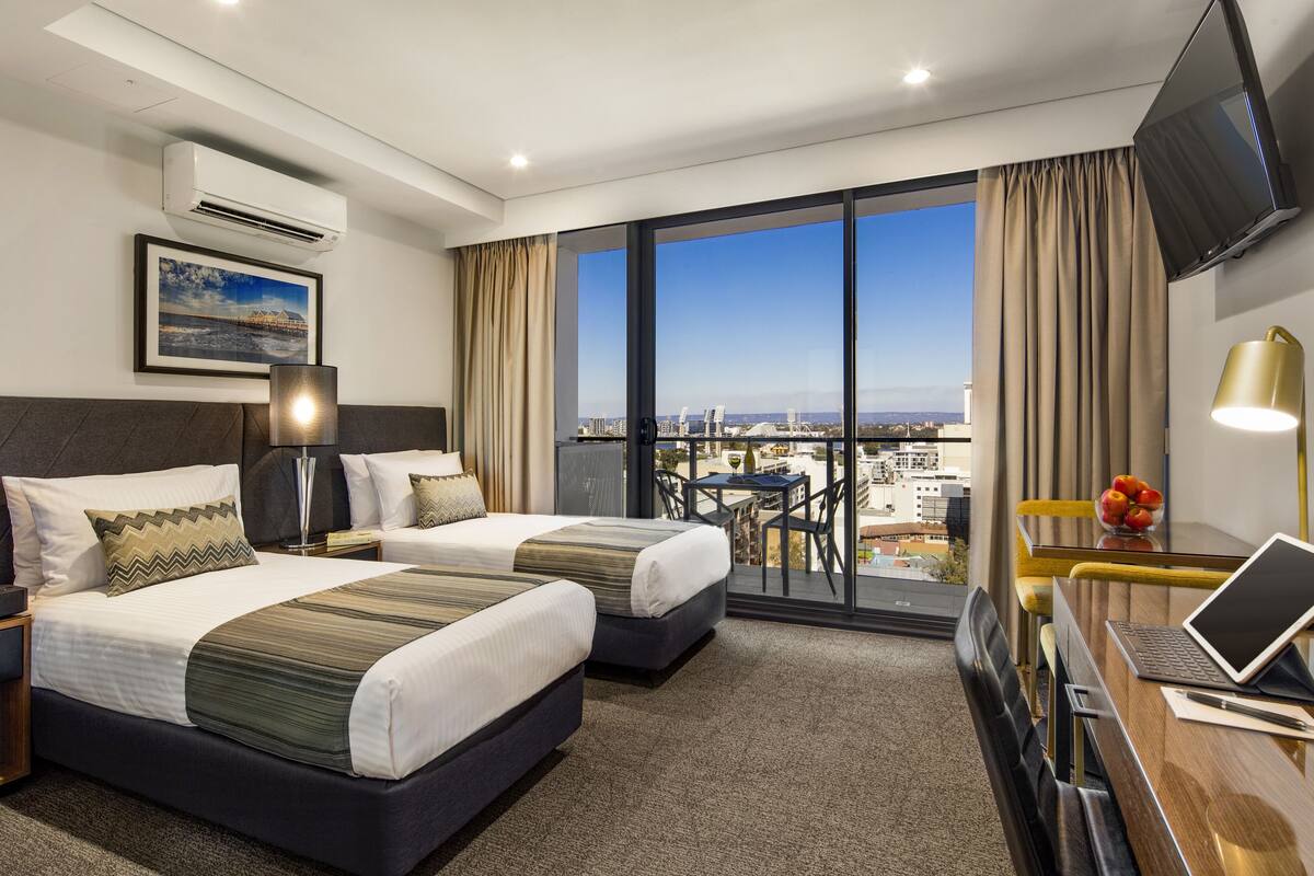A hotel room with two beds, a desk with a laptop, a TV, and a balcony with a city view.