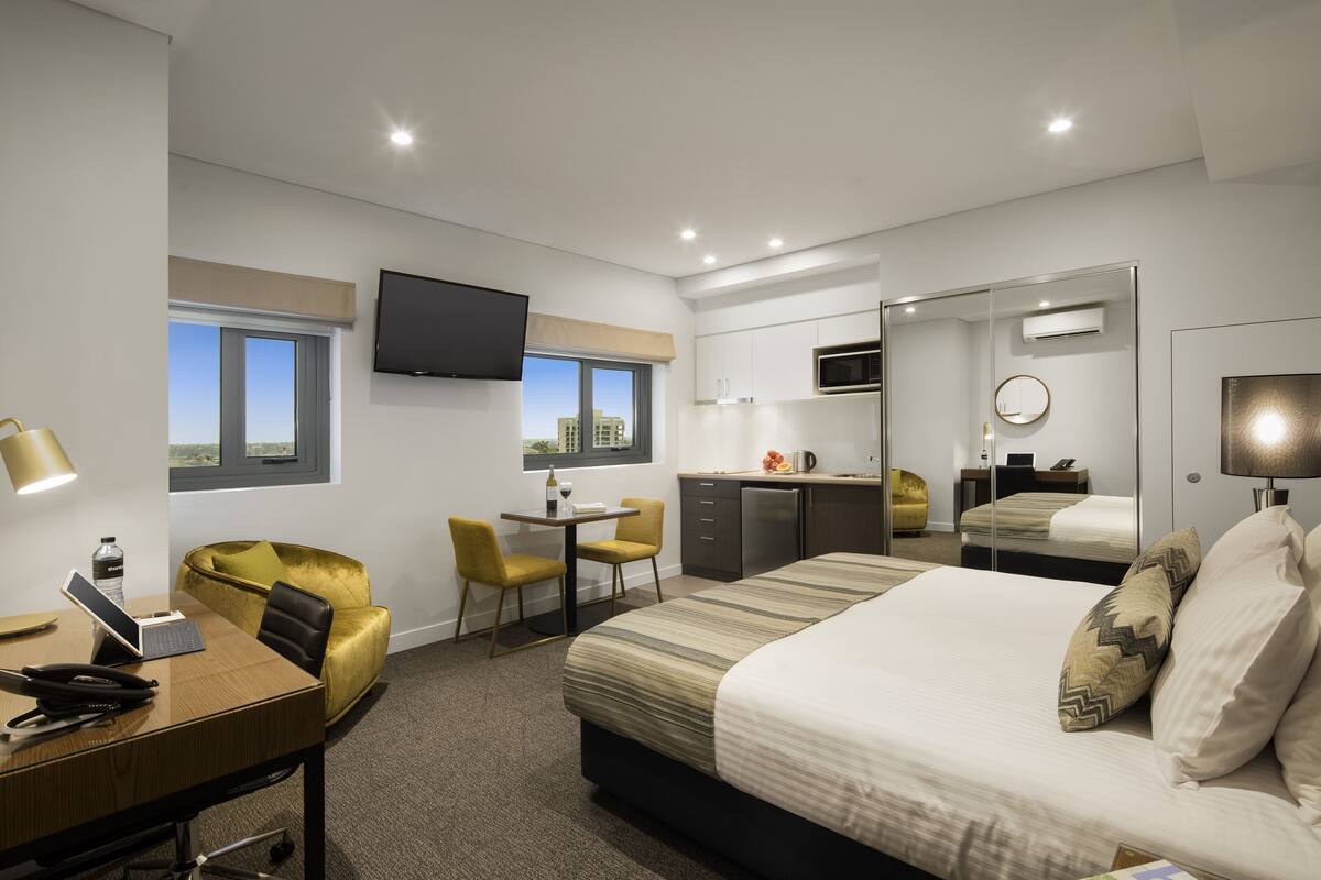 A modern hotel room with a bed, desk, chairs, a TV, a kitchenette, and a window with a view.
