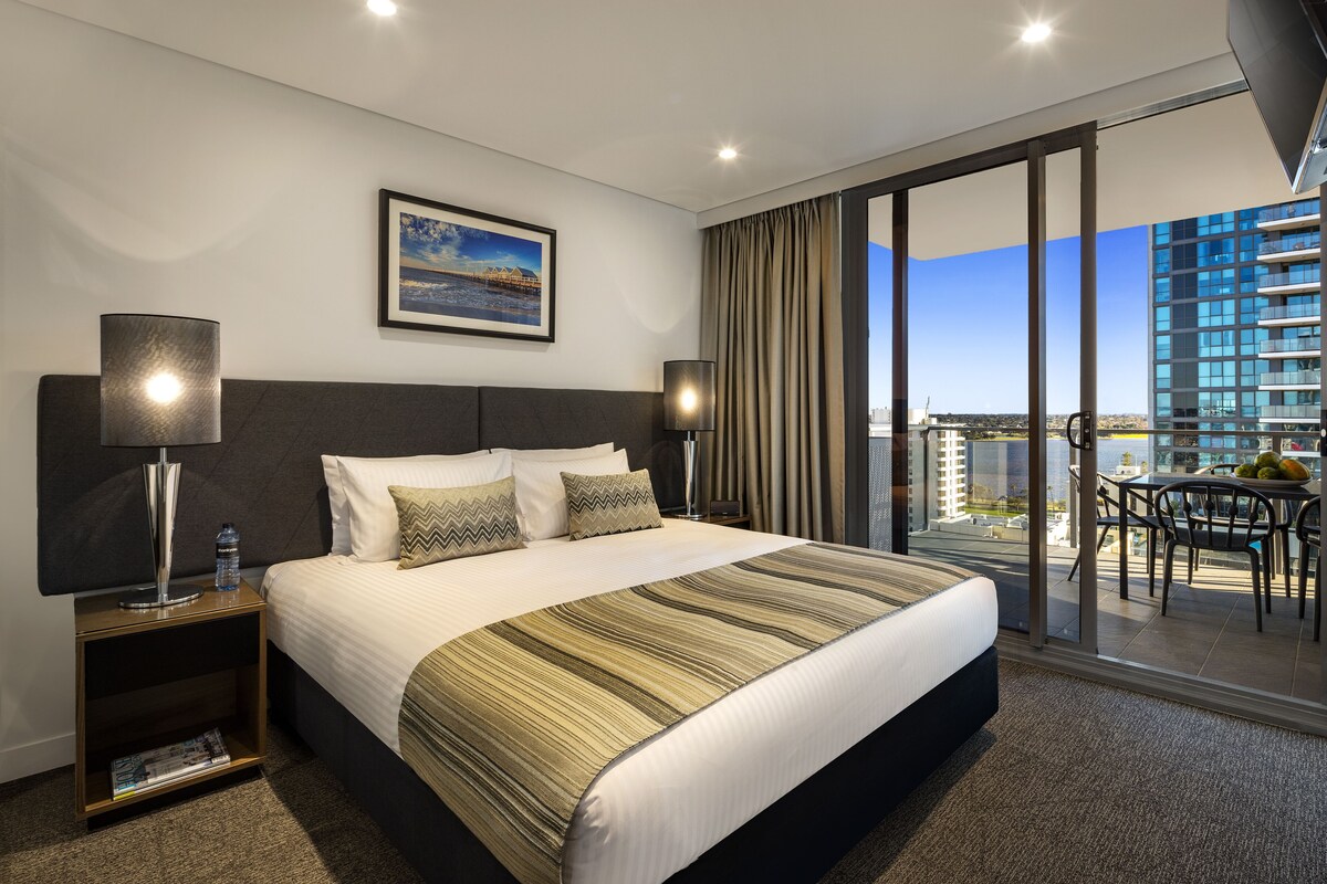 Habitación de hotel con una cama grande, una mesita de noche con lámpara, un cuadro en la pared y un balcón con mesa y sillas.