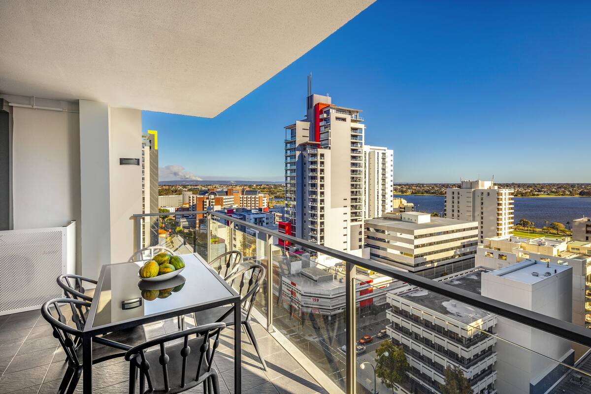 A balcony with a table and chairs, overlooking a cityscape with buildings and a body of water.