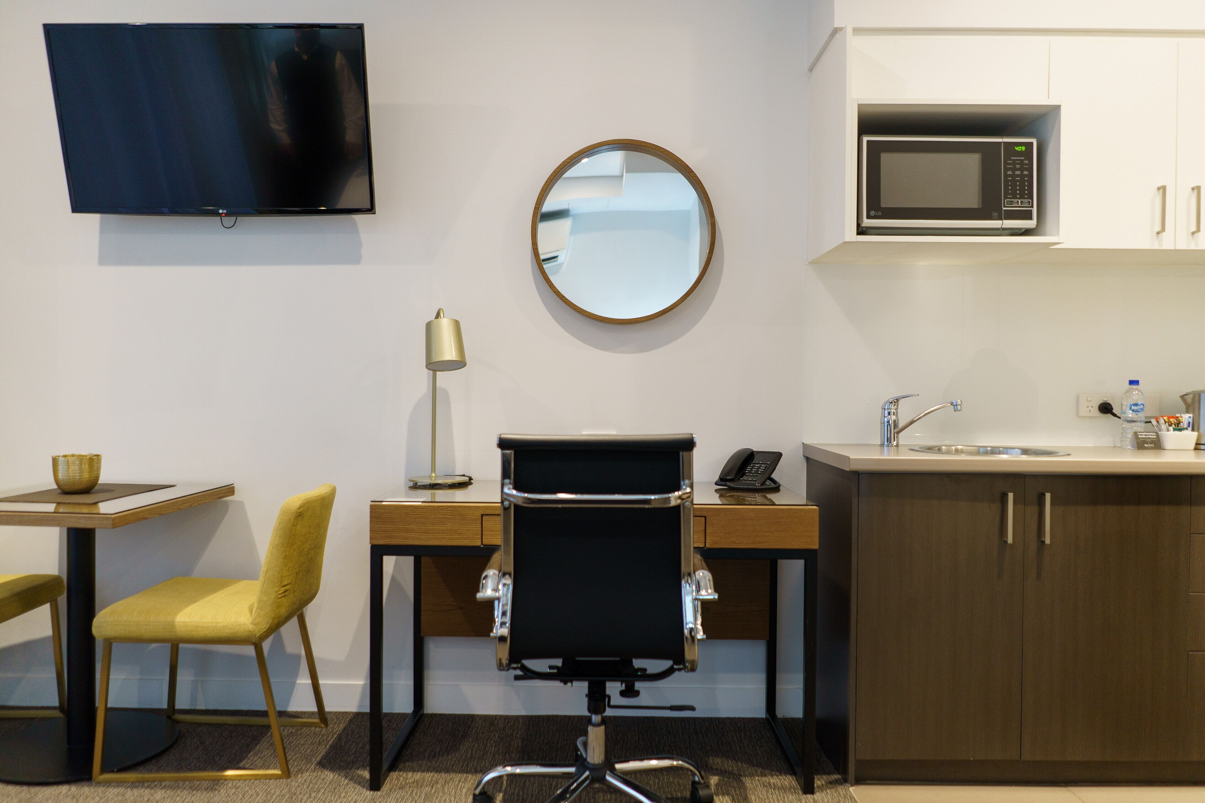 A hotel room with a flat-screen TV, a round mirror, a microwave, a desk with a chair, a sink, and a small table with a bowl.