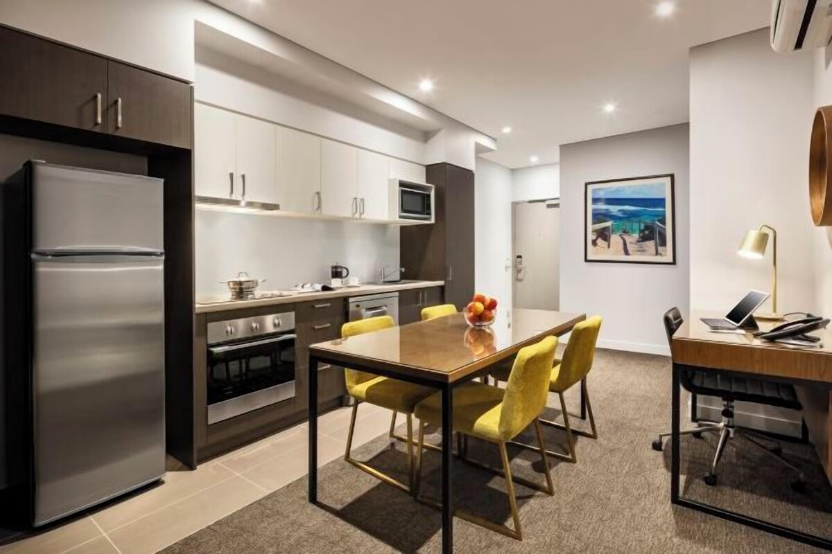 A modern kitchen with a stainless steel refrigerator, microwave, and oven. There is a dining table with yellow chairs and a fruit bowl.