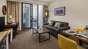 A modern hotel room with a sofa, a coffee table, a desk with a laptop, a dining table with oranges and a water bottle, and a large window with a view of buildings. - Quest East Perth (East Perth)