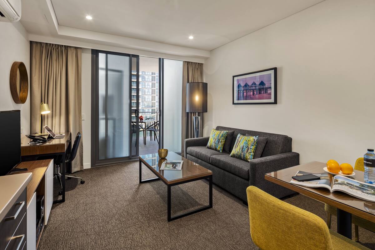 A modern hotel room with a sofa, a coffee table, a desk with a laptop, a dining table with oranges and a water bottle, and a large window with a view of buildings.