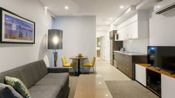 A modern living room with a sofa, coffee table, and a dining area with chairs. There is a kitchen with cabinets and a TV on a stand.