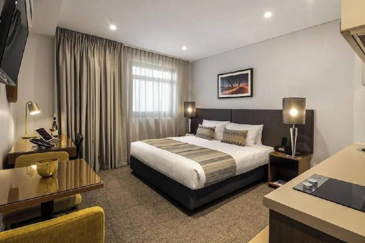 A hotel room with a bed, bedside tables, a desk, a chair, a TV, and a window with curtains.